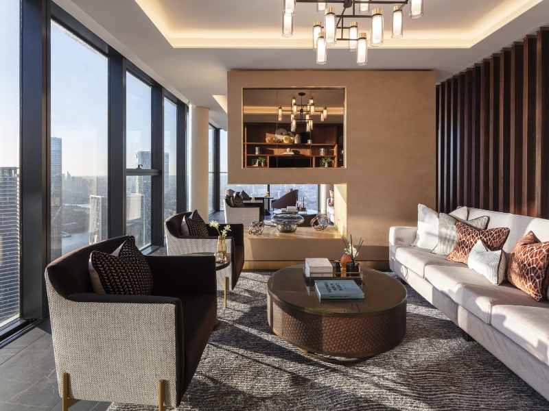 Interior photo of a sophisticated lounge area for residents, with plush long sofa with cushions and armchairs, tables with a view through floor to ceiling windows across Canary Wharf