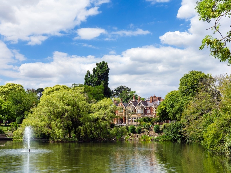 Exterior photo of the grounds at Bletchley Park, surrounded by mature trees, with a lake with fountain in front