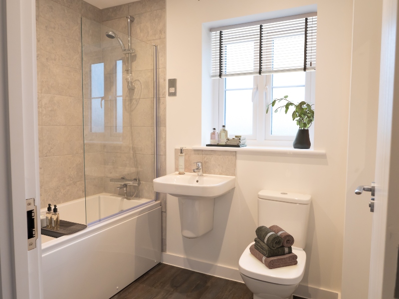 Interior view of a CGI dressed bathroom taken in an actual 4 bed house at Benson Grange, showing bath with glass screen, handbasin and toilet with window behind