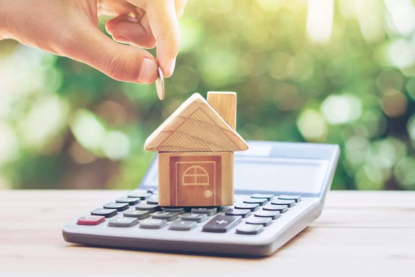 Model of a house on top of a calculator, with a hand holding a coin above it.