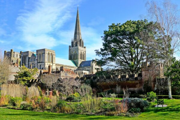 Chichester Cathedral in the background taken from the surrounding gardens with thick green trees and bushes on a clear summer day.