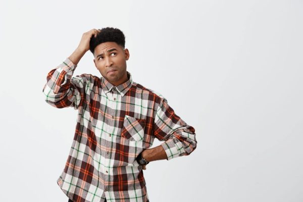 Man stood scratching head appearing confused in a grey and red check long-sleeved shirt.
