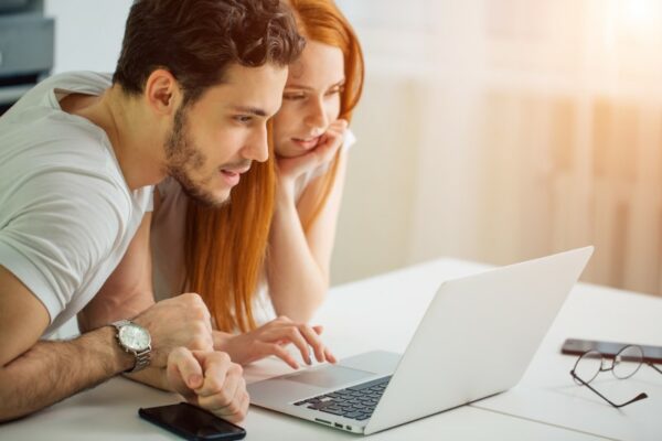 Two people looking excitedly at a laptop in their home doing some research with some sunshine coming through the window behind them.